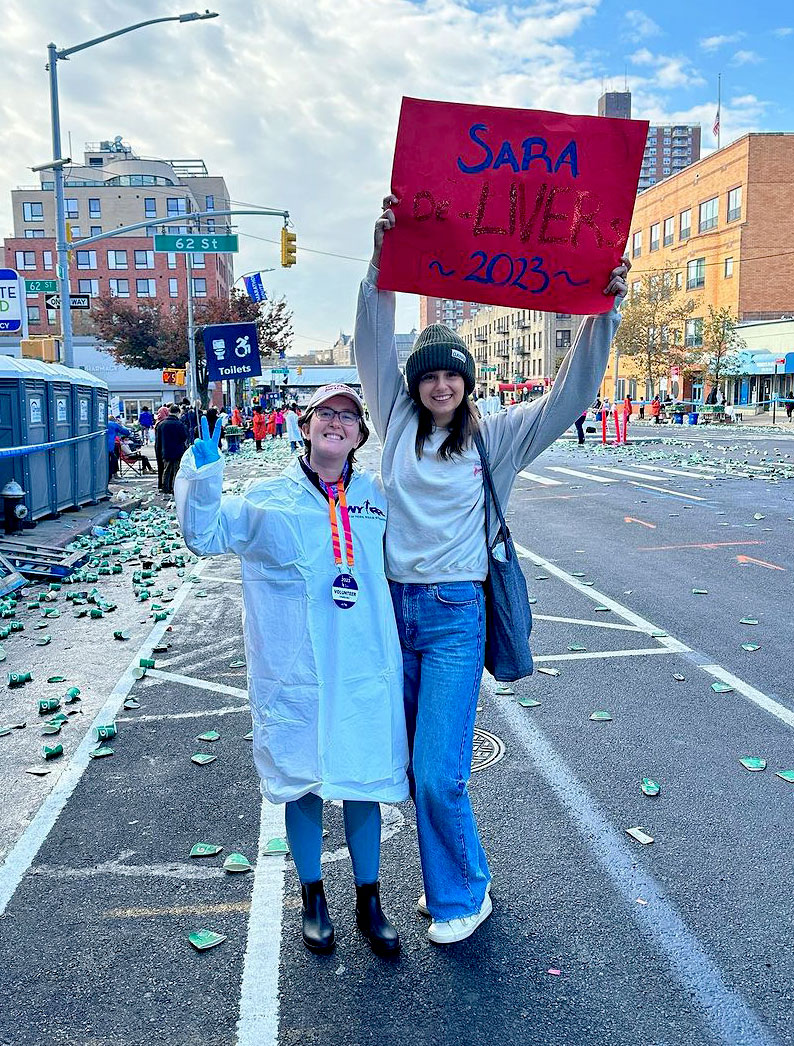 Zoe and Vicky standing in street littered with cups following race. Holding a poster that reads, Sara De-Livers 2023
